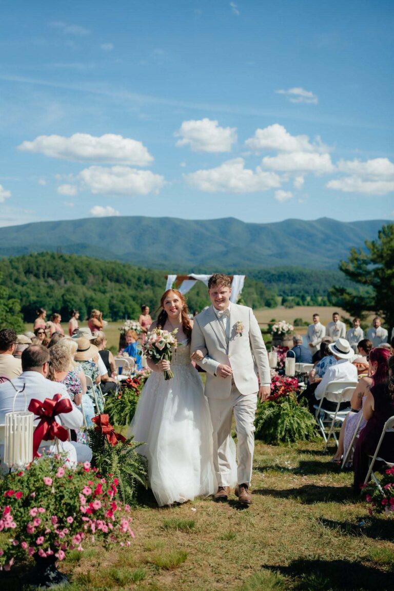 Newlyweds walking back down a flower-lined aisle past seated guests with the mountains behind