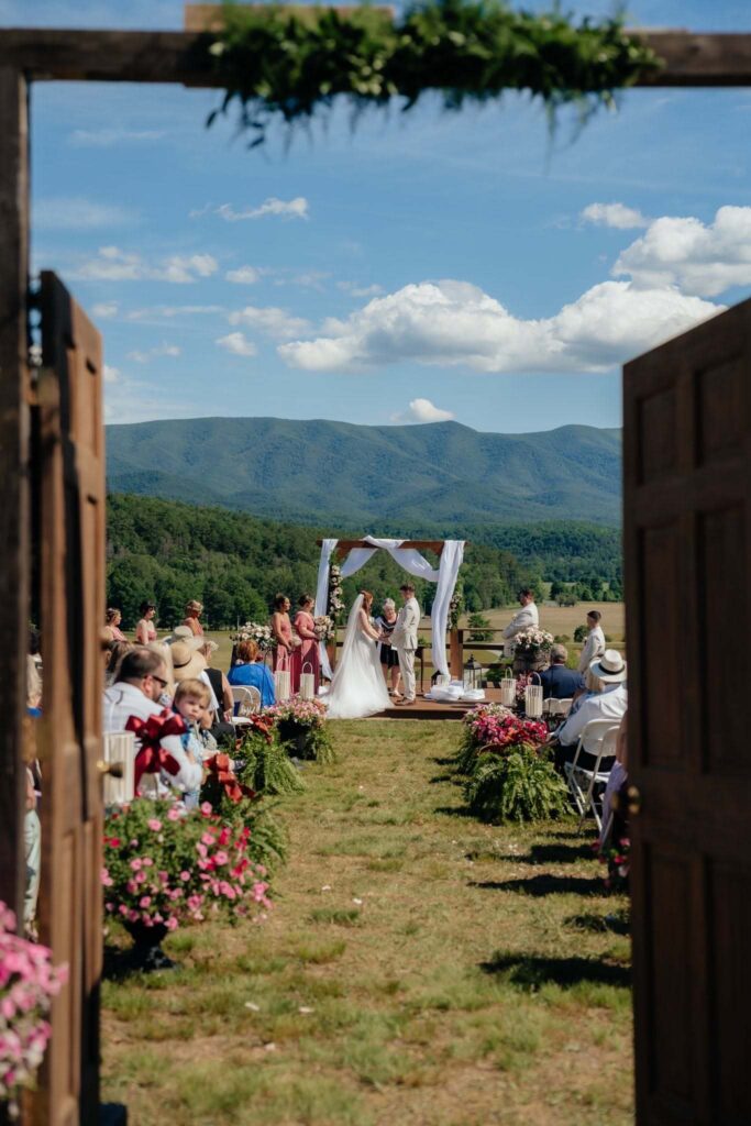 View through open barn doors to the ceremony arch, seated guests and mountain ridge beyond