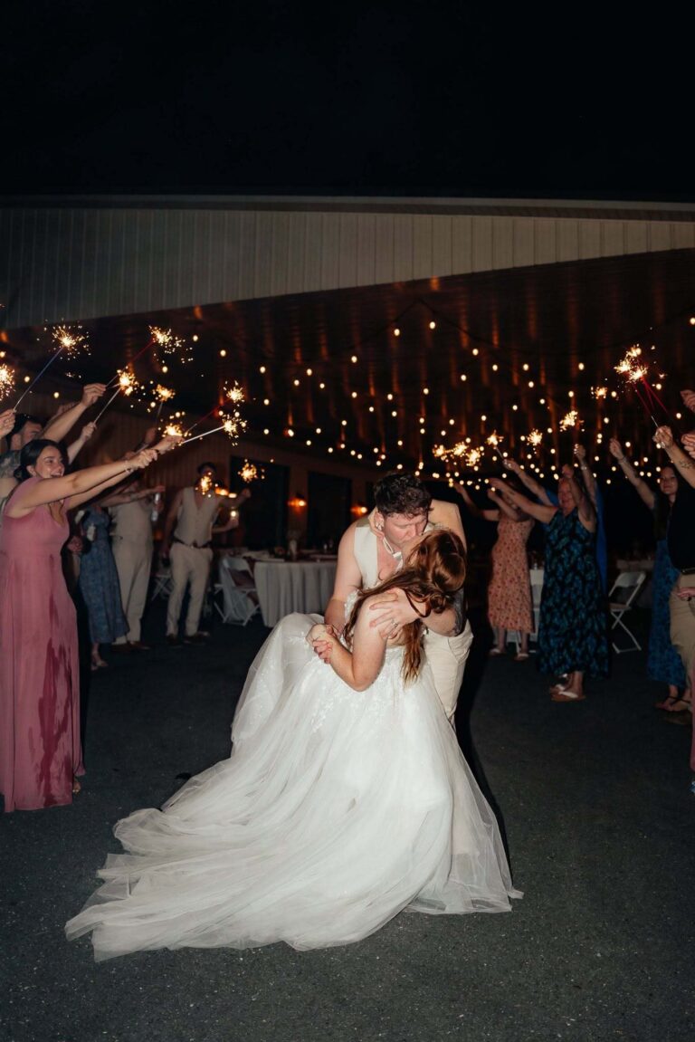 Bride and groom dancing under sparklers held by guests in the string-lit waterside pavilion