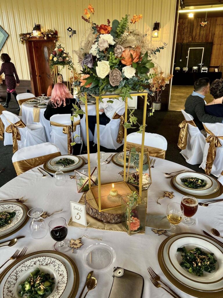 Round reception table set with white linens, gold accents and a tall floral centerpiece
